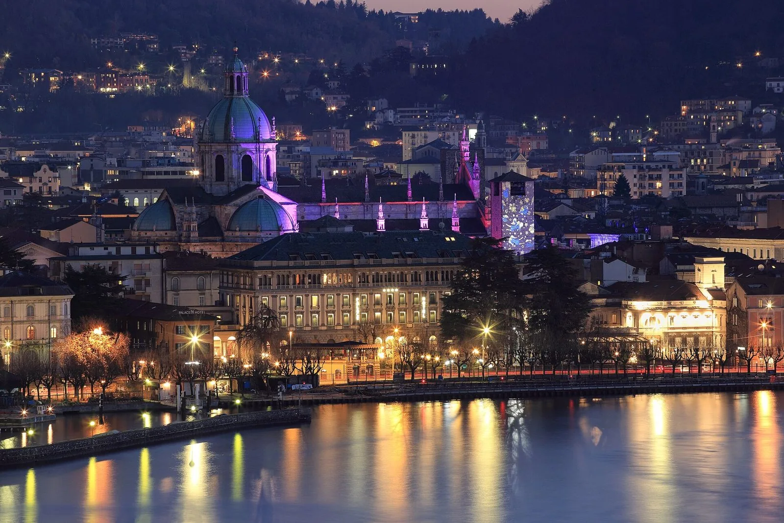Il centro di Como di sera durante la Città dei Balocchi: la cupola del Duomo illuminata di viola e la torre del Broletto con proiezioni natalizie, con le luci riflesse nel lago
