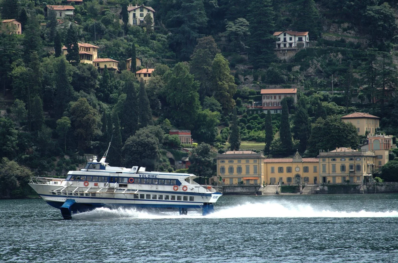 Un aliscafo bianco e blu naviga veloce sul lago di Como davanti a una villa gialla e alla collina boscosa punteggiata di case