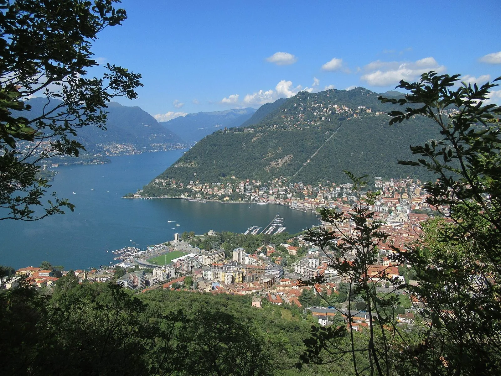 Veduta dall'alto della città di Como e del primo bacino del lago, con la collina di Brunate e le montagne sullo sfondo in una giornata di sole