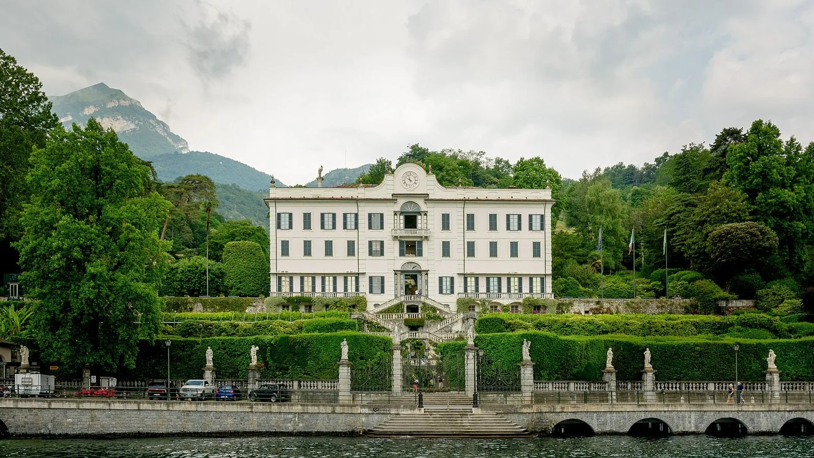 Villa Carlotta a Tremezzo vista dal lago, con la facciata bianca, la scalinata, le siepi e le statue sulla balaustra a riva, colline boscose sullo sfondo